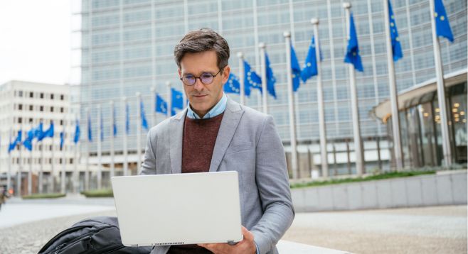 Businessman working on a laptop in front of the European Commission building with several EU flags in the background